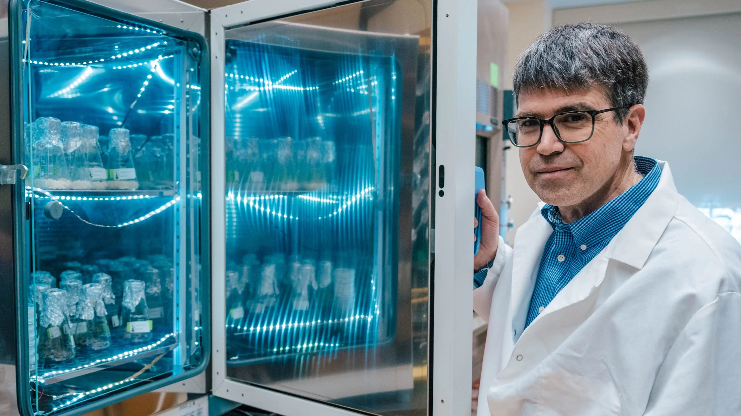 A researcher in a white lab coat opens a glowing blue incubator filled with labeled sample flasks in a laboratory.