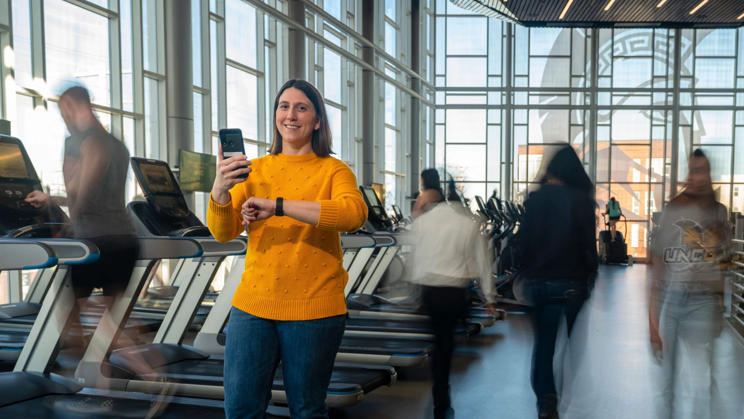 A woman in a yellow sweater holds a smartphone and checks her smartwatch while standing in a busy university fitness center.